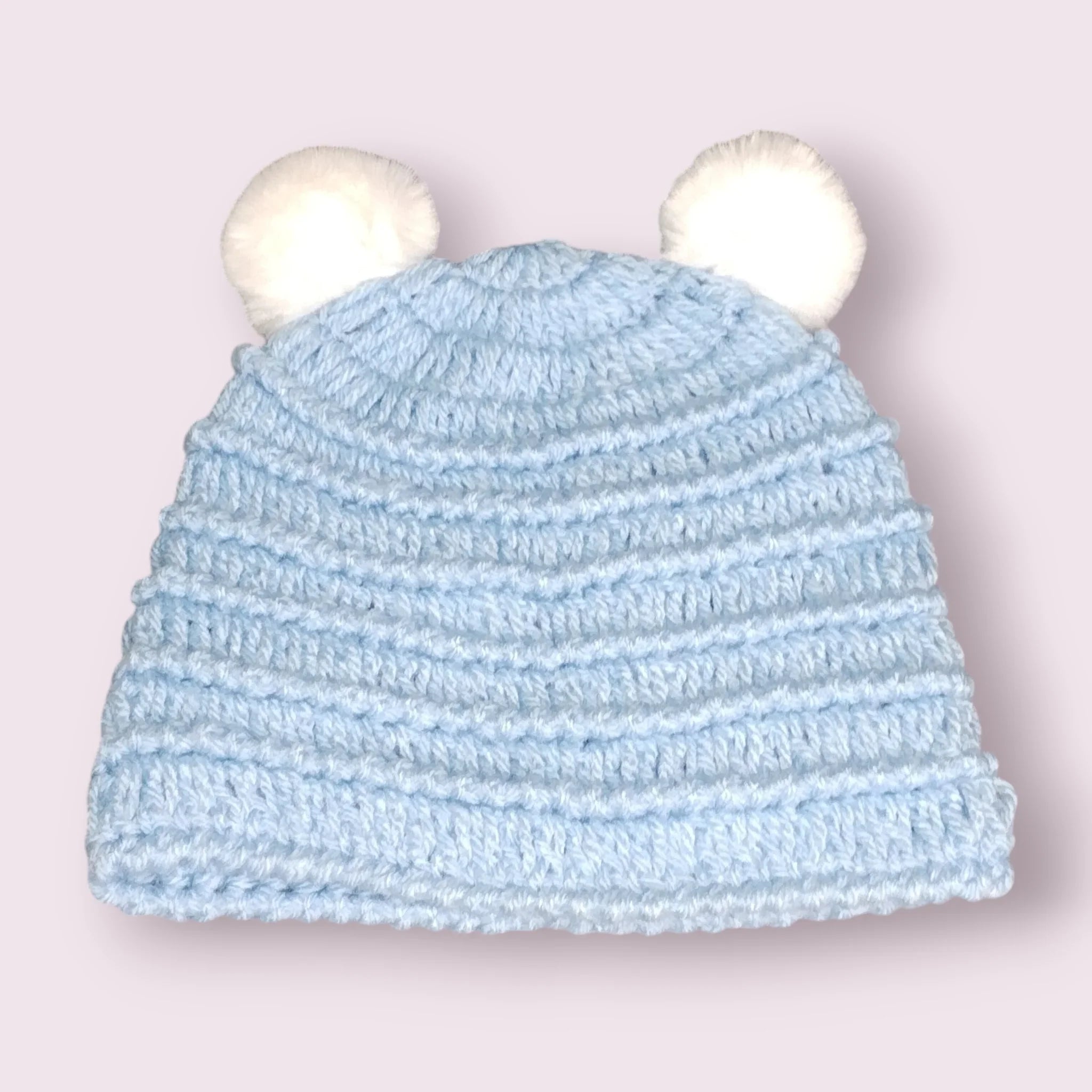Handmade light blue crochet baby hat with two white pom pom ears on a soft pink background
