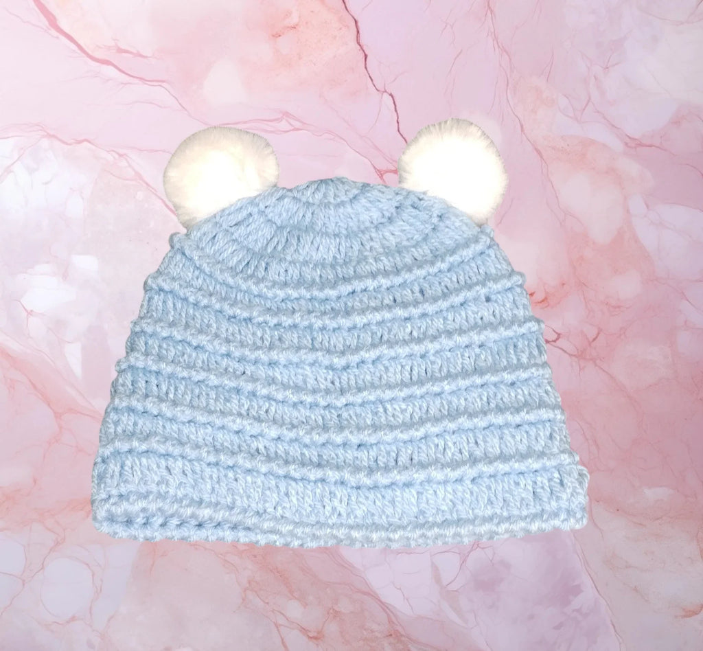 Handmade light blue crochet baby hat with white pom-pom ears on a pink marble background