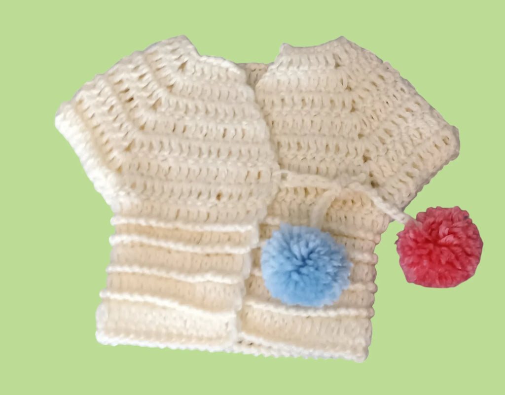 Handmade cream crochet baby vest with blue and red pom-poms on green background