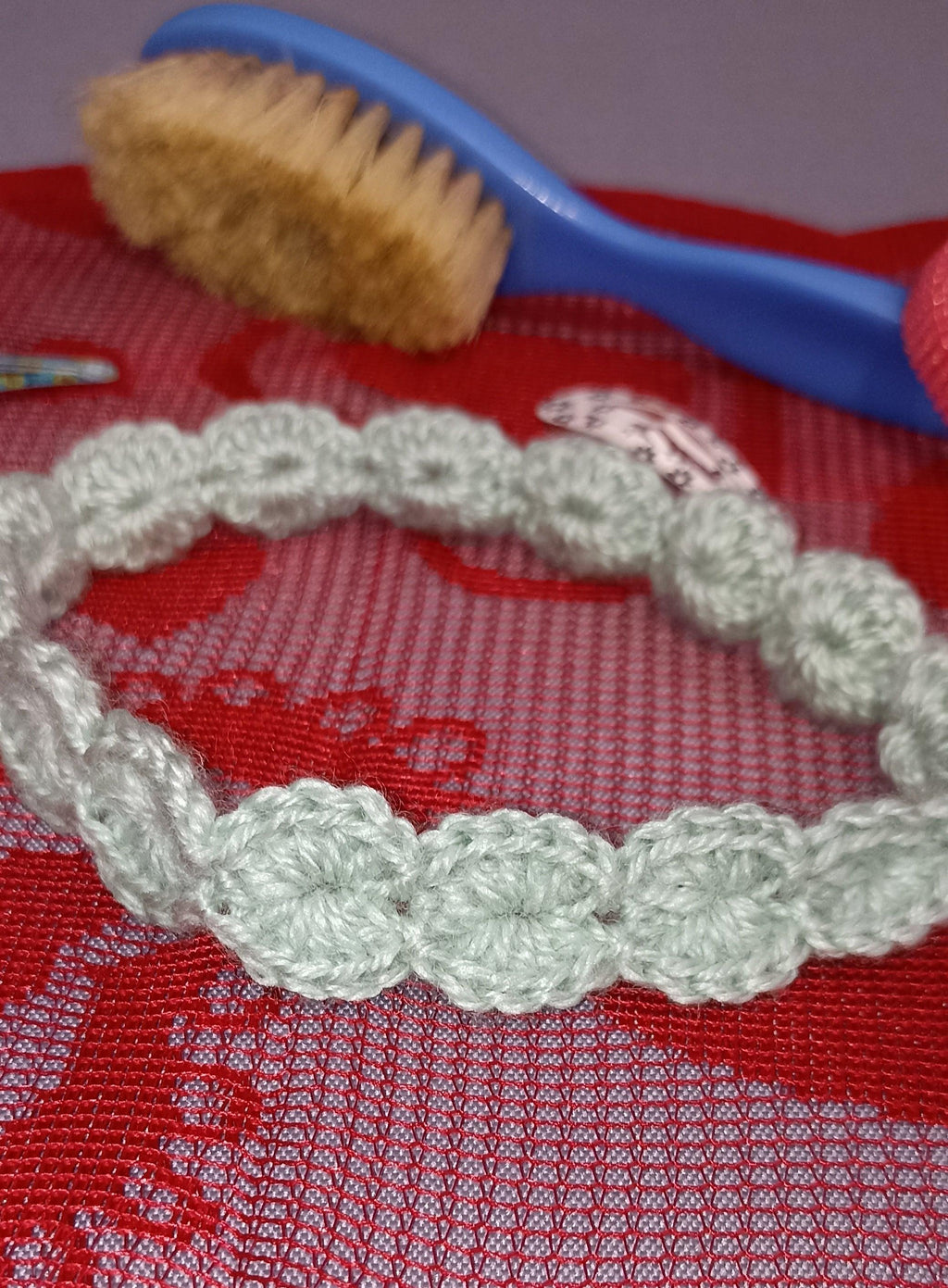 Handmade crochet wool headband in light green, displayed on red fabric near a baby brush