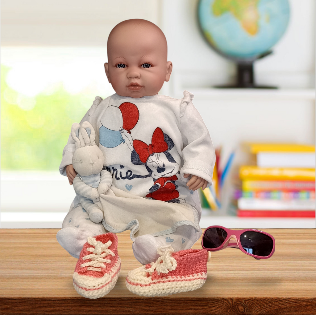 Handmade crochet baby shoes in pink and white by Croch'pelotes by Sophie, displayed with a baby doll, plush bunny, and kids' sunglasses on a wooden surface, with books and a globe in the background.