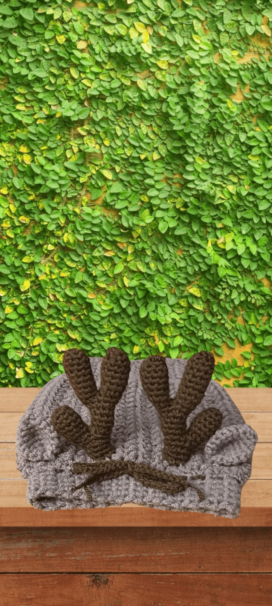 Handmade crochet grey snood with brown antlers on wooden table, green leafy wall background